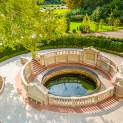 Luftaufnahme der Donauquelle in Donaueschingen, ein rundes Steinbecken mit verzierter Balustrade und Skulpturen, umgeben von einem Park.