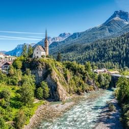 White church with tall spire on rocky outcrop above turquoise river, surrounded by alpine village, forests, and dramatic mountain peaks.