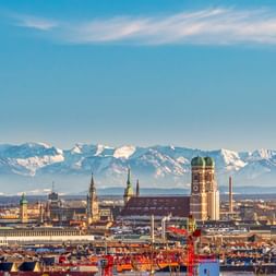 Münchner Stadtbild mit Frauenkirche-Türmen und historischen Gebäuden, schneebedeckte Alpen am Horizont unter blauem Himmel.