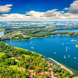 Luftaufnahme vom Lötzener See in Masuren mit Segelbooten, bewaldeten Inseln und grünen Ufern unter blauem Himmel mit weißen Wolken.