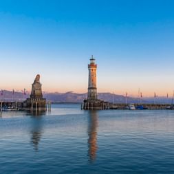 Lindauer Hafeneinfahrt mit Leuchtturm und bayerischem Löwen auf Steinsockeln im Bodensee bei Sonnenuntergang, Alpen im Hintergrund.
