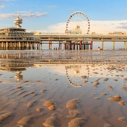 Scheveninger Pier mit rundem Gebäude und Riesenrad, gespiegelt im nassen Sand bei Ebbe. Der Strand zeigt verstreute Steine und Fußspuren.