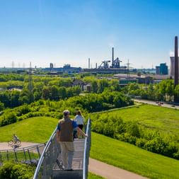 Erhöhter Steg im Landschaftspark Duisburg mit Besuchern, Blick auf grüne Parkfläche, Radfahrer auf Weg unten und Industrieanlagen im Hintergrund.