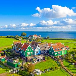 Luftaufnahme traditioneller grüner Holzhäuser mit roten Ziegeldächern im Dorf Marken, umgeben von grünen Wiesen und dem blauen IJsselmeer.