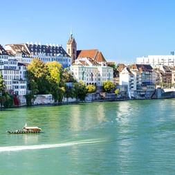 Panoramablick auf Basel mit dem türkisfarbenen Rhein, Booten, historischen Gebäuden am Ufer, einer Brücke und baumgesäumter Promenade.