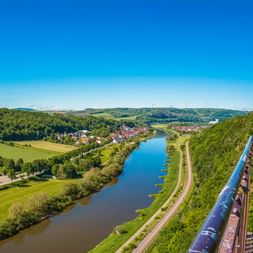 Panoramablick vom Weser Skywalk bei Beverungen auf die Weser, die sich durch grünes Tal mit Wäldern, Wiesen und einer Stadt schlängelt.