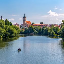 Blick auf Nürtingen am Neckar mit Kirchturm, historischen Gebäuden und einem kleinen Boot auf dem Wasser, eingerahmt von grünen Bäumen.