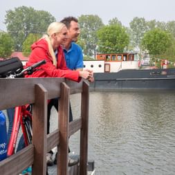 Zwei Radfahrer mit Fahrrädern an Holzgeländer mit Blick auf Kanal und Schiff Magnifique II in den Niederlanden.