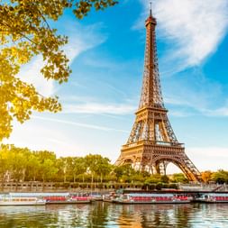 The Eiffel Tower rises above the Seine River in Paris on a sunny day. Tour boats are moored along the riverbank, framed by green trees.