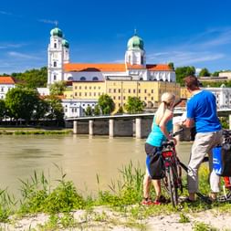 Zwei Radfahrer mit Packtaschen stehen am Inn und blicken auf Passaus Altstadt mit Domtürmen und Brücke unter blauem Himmel.