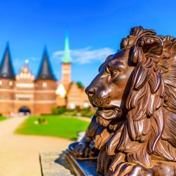 Bronzelöwenstatue im Profil mit dem historischen Holstentor in Lübeck im Hintergrund unter blauem Himmel.