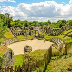 Ancient Roman amphitheater ruins in Saintes with grass-covered arena, stone seating tiers, and wooden fence. Church tower visible in background under cloudy sky.