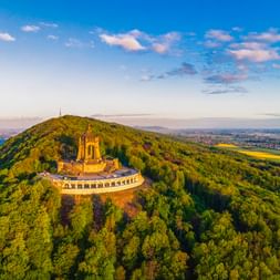 Luftaufnahme des Kaiser-Wilhelm-Denkmals auf einem bewaldeten Hügel in Porta Westfalica mit umliegender Landschaft und blauem Himmel.
