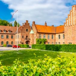 Backsteingebäude des Klosters in Roskilde mit Treppengiebeln und orangefarbenen Ziegeldächern, umgeben von grünem Rasen und Hecken.
