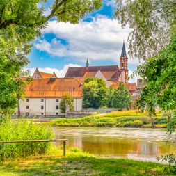 Blick auf die Karmelitenkirche mit rotem Backsteinturm und historische Gebäude an der Donau in Straubing, eingerahmt von grünen Bäumen.