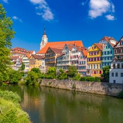 Historische bunte Fachwerkhäuser am Neckar in Tübingen mit Kirchturm, Steinmauer und grünem Flussufer unter blauem Himmel.