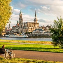 Fahrrad auf Weg am Elbufer mit Dresdner Altstadtsilhouette mit Kirchtürmen und Barockgebäuden auf der anderen Flussseite unter bewölktem Himmel.
