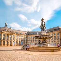 Place de la Bourse in Bordeaux mit prächtigem Brunnen in der Mitte, klassischer französischer Architektur und blauem Himmel mit Wolken.