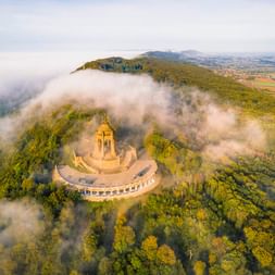 Luftaufnahme des Kaiser-Wilhelm-Denkmals auf bewaldetem Hügel in Porta Westfalica, umgeben von Morgennebel mit Landschaft im Hintergrund.