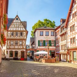 Kopfsteinpflasterplatz in Mainz, umgeben von bunten Fachwerkhäusern. Ein Café mit Außenbestuhlung befindet sich in der Mitte unter blauem Himmel.