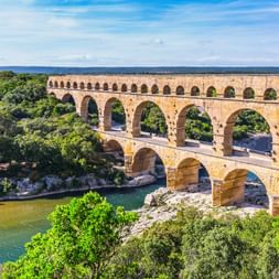 Pont du Gard, antikes römisches Aquädukt mit drei Bogenreihen über dem Fluss Gardon, umgeben von üppiger grüner Vegetation unter blauem Himmel.