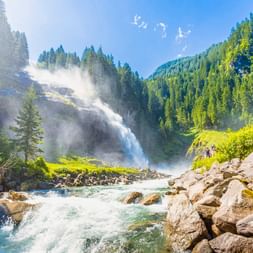 Krimml Waterfalls cascading down forested mountainside with mist, rocky stream in foreground, surrounded by green coniferous forests.