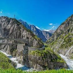 Historic stone bridge spanning a deep gorge near Andermatt with rushing river below, steep rocky cliffs, and snow-capped mountains in background.
