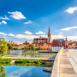 Steinerne Brücke über die Donau zur Regensburger Altstadt mit Domturm, bunten historischen Gebäuden und blauem Himmel.