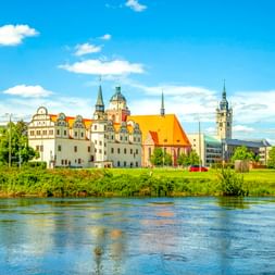 Dessauer Stadtschloss mit weißer Fassade und Türmen an der Elbe, mit Kirchtürmen und modernen Gebäuden unter blauem Himmel.