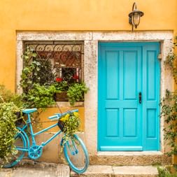 Blaues Fahrrad mit Pflanzen geschmückt, das an einer gelben Wand neben türkisfarbener Tür und Fenster mit Pflanzen in Rovinj, Kroatien lehnt.