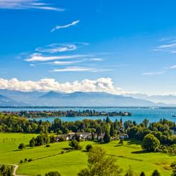 Luftaufnahme vom Bodensee bei Lindau mit grünen Wiesen, Bäumen und einer Insel. Berge und weiße Wolken im Hintergrund sichtbar.