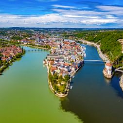 Luftaufnahme von Passau mit der historischen Altstadt auf einer Halbinsel, wo zwei Flüsse zusammenfließen, umgeben von grünen Wäldern und Brücken.