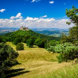 Burg Hohenzollern thront auf einem bewaldeten Hügel in Süddeutschland, umgeben von grünen Wiesen und Bäumen unter blauem Himmel mit weißen Wolken.