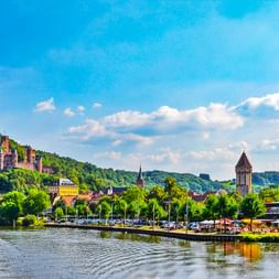 Blick auf Wertheim am Main mit Burgruine auf grünem Hügel, Kirchturm und Uferpromenade unter blauem Himmel.