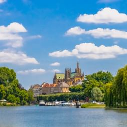 Metzer Kathedrale über historischen Gebäuden, vom Fluss aus gesehen mit grünen Bäumen und Weidenzweigen unter blauem Himmel mit weißen Wolken.