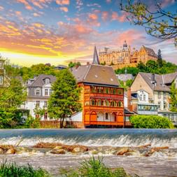 Marburger Landgrafenschloss auf Hügel über bunten historischen Häusern entlang der Lahn mit Wehr bei Sonnenuntergang unter dramatischem Himmel.