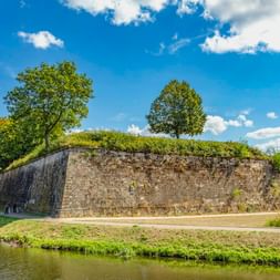 Historische Steinmauer der Festung Saarlouis mit grasbewachsenen Wällen und Bäumen, umgeben von einem Wassergraben unter blauem Himmel.