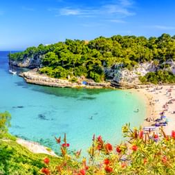 Strand Cala Llombards mit türkisfarbenem Wasser, umgeben von Felsklippen und grüner Vegetation. Badegäste entspannen am Sandstrand unter blauem Himmel.