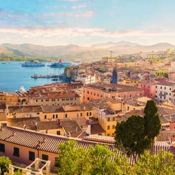 Luftaufnahme des Hafens von Portoferraio mit bunten Gebäuden, Terrakottadächern, Fähren in der Bucht und Bergen im Hintergrund bei Sonnenuntergang.