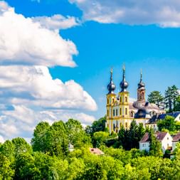 Wallfahrtskirche Käppele mit gelben Barocktürmen und Zwiebeltürmen auf einem grünen Hügel in Würzburg, umgeben von Bäumen unter blauem Himmel.