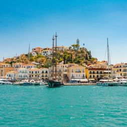 Türkisfarbenes Hafenwasser der Insel Poros mit Segelbooten und Yachten. Bunte Gebäude säumen die Uferpromenade unter einer Hangstadt mit Uhrturm.