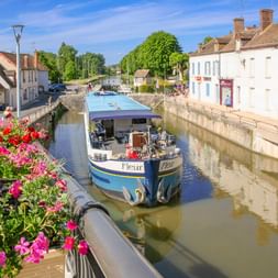 MS Fleur Kreuzfahrtschiff mit blauem Dach in einem schmalen Kanal in Frankreich, umgeben von bunten Blumen und historischen Gebäuden.