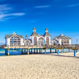 Historische Seebrücke Sellin in der Ostsee mit weißem Pavillon mit Zwillingstürmen, Sandstrand im Vordergrund unter blauem Himmel.