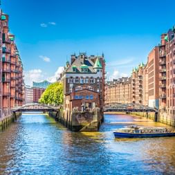Touristenboot auf Kanal in Hamburgs Speicherstadt mit roten Backsteingebäuden und grünen Kupferdächern. Blauer Himmel und Brücken im Hintergrund.
