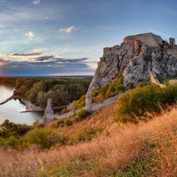 Ruinen der Burg Devin auf einem Felsen mit Blick auf die Donau bei Sonnenuntergang, mit goldenem Gras im Vordergrund und blauem Himmel.