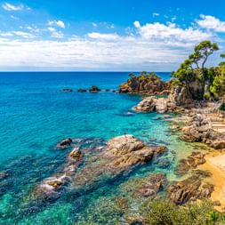 Holzsteg schlängelt sich entlang felsiger spanischer Küste mit türkisfarbenem Wasser, kleinem Strand und Pinien auf Klippen.