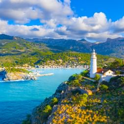 Weißer Leuchtturm auf felsiger Klippe mit Blick auf die Bucht von Port de Sóller auf Mallorca, mit türkisfarbenem Wasser, Küstenort und Tramuntana-Bergen.