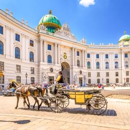 Gelber Fiaker vor der Alten Hofburg in Wien mit weißer Barockfassade und grünen Kupferkuppeln unter blauem Himmel.