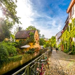 Kopfsteinpflasterstraße in der Freiburger Altstadt mit Kanal, efeubedeckten Gebäuden, Fahrrädern und gelbem Haus unter blauem Himmel.