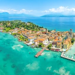 Luftaufnahme der Halbinsel Sirmione am Gardasee mit türkisfarbenem Wasser, historischen Gebäuden, Burg und Booten. Berge im Hintergrund.